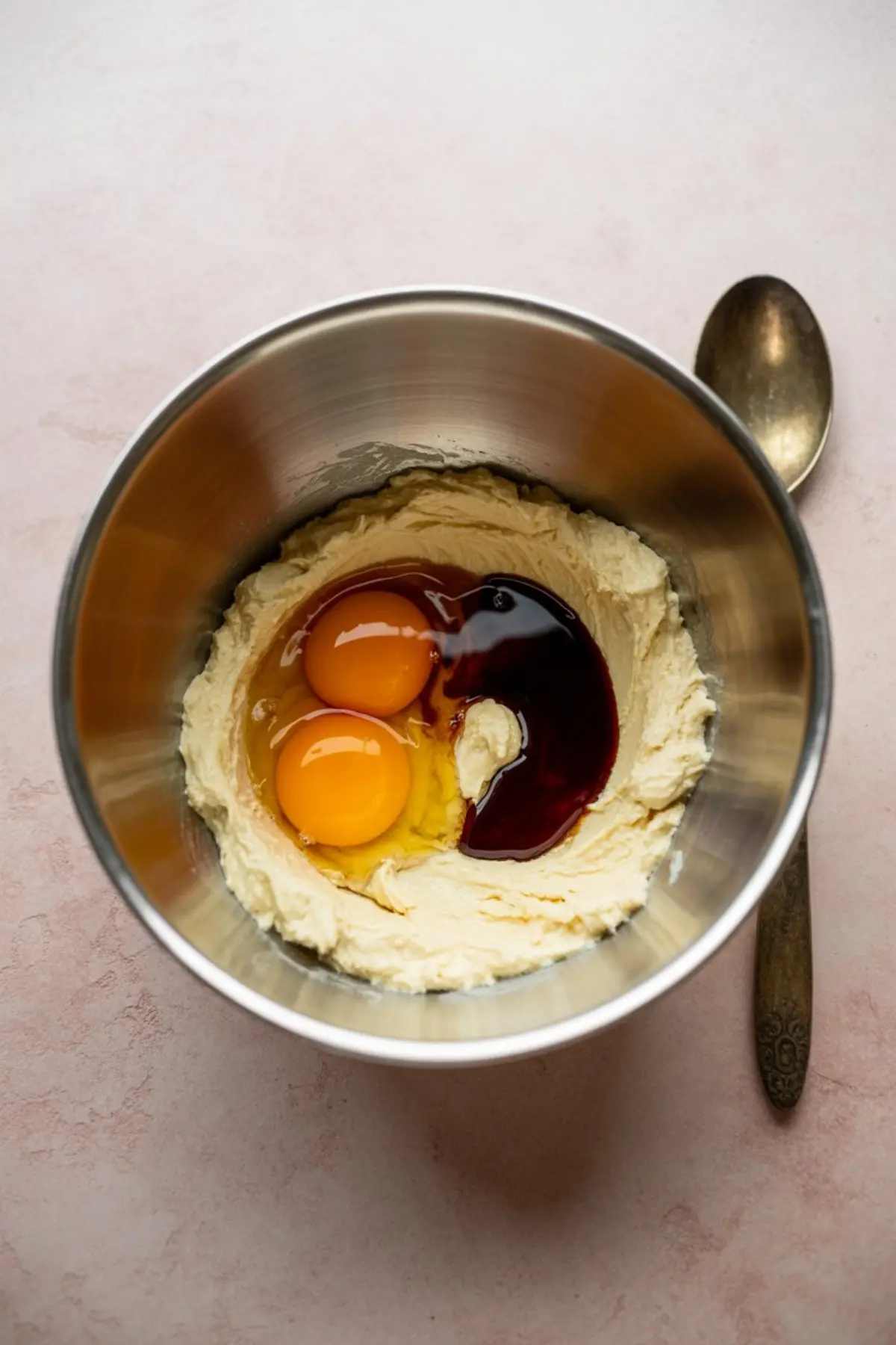 Two raw eggs and dark vanilla extract added to creamed butter mixture in a metal bowl, ready to be blended for cake batter.
