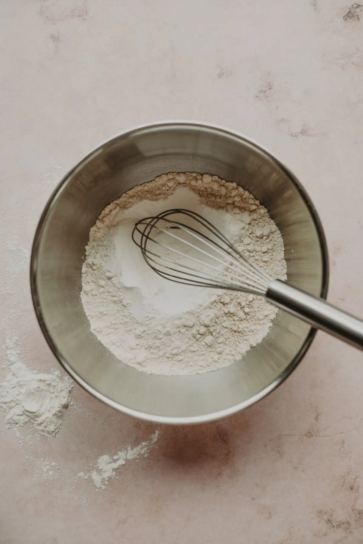 Stainless steel mixing bowl filled with all-purpose flour, baking powder, and a metal whisk, set on a light pink surface with scattered flour for a cookie baking scene.