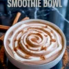 Cinnamon roll smoothie bowl styled with spiraled yogurt drizzle and sprinkled cinnamon powder, placed on a dark marble surface with cinnamon sticks.