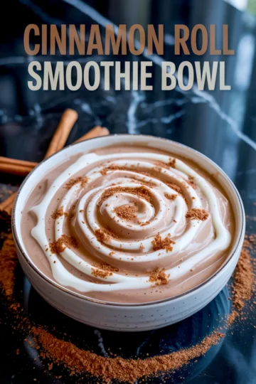 Cinnamon roll smoothie bowl styled with spiraled yogurt drizzle and sprinkled cinnamon powder, placed on a dark marble surface with cinnamon sticks.