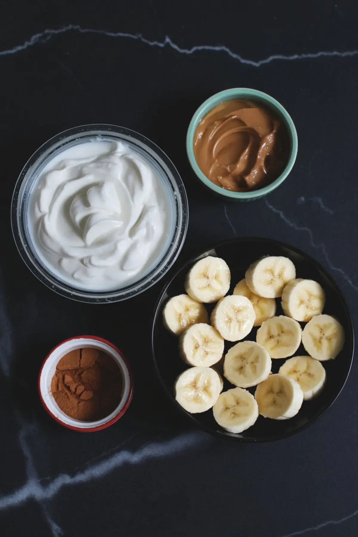 Flat lay of cinnamon banana smoothie ingredients including Greek yogurt, creamy peanut butter, banana slices, and ground cinnamon in separate bowls.
