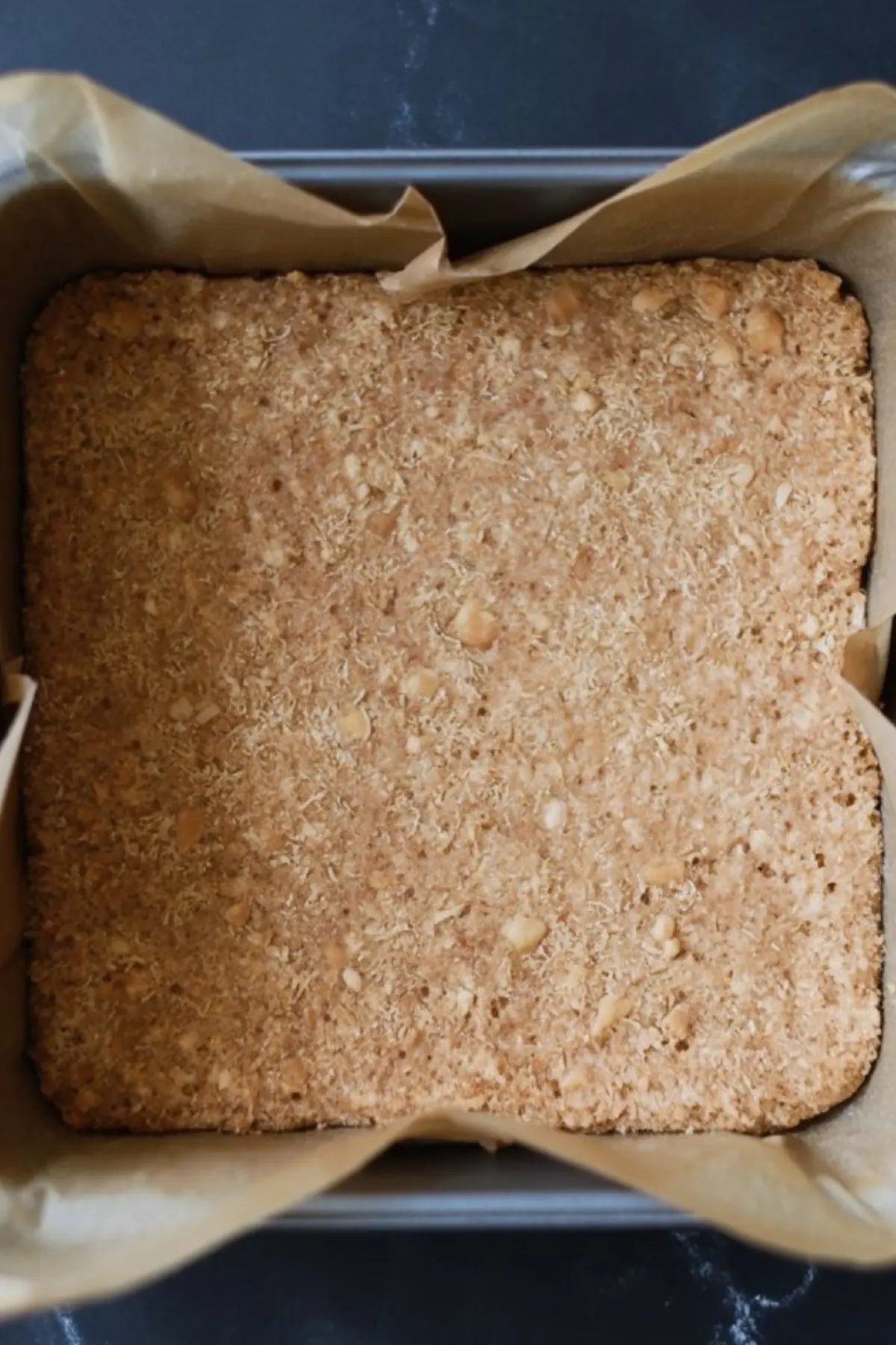 Pressed graham cracker coconut crust in a parchment-lined baking pan, ready to be baked for layered dessert bars.