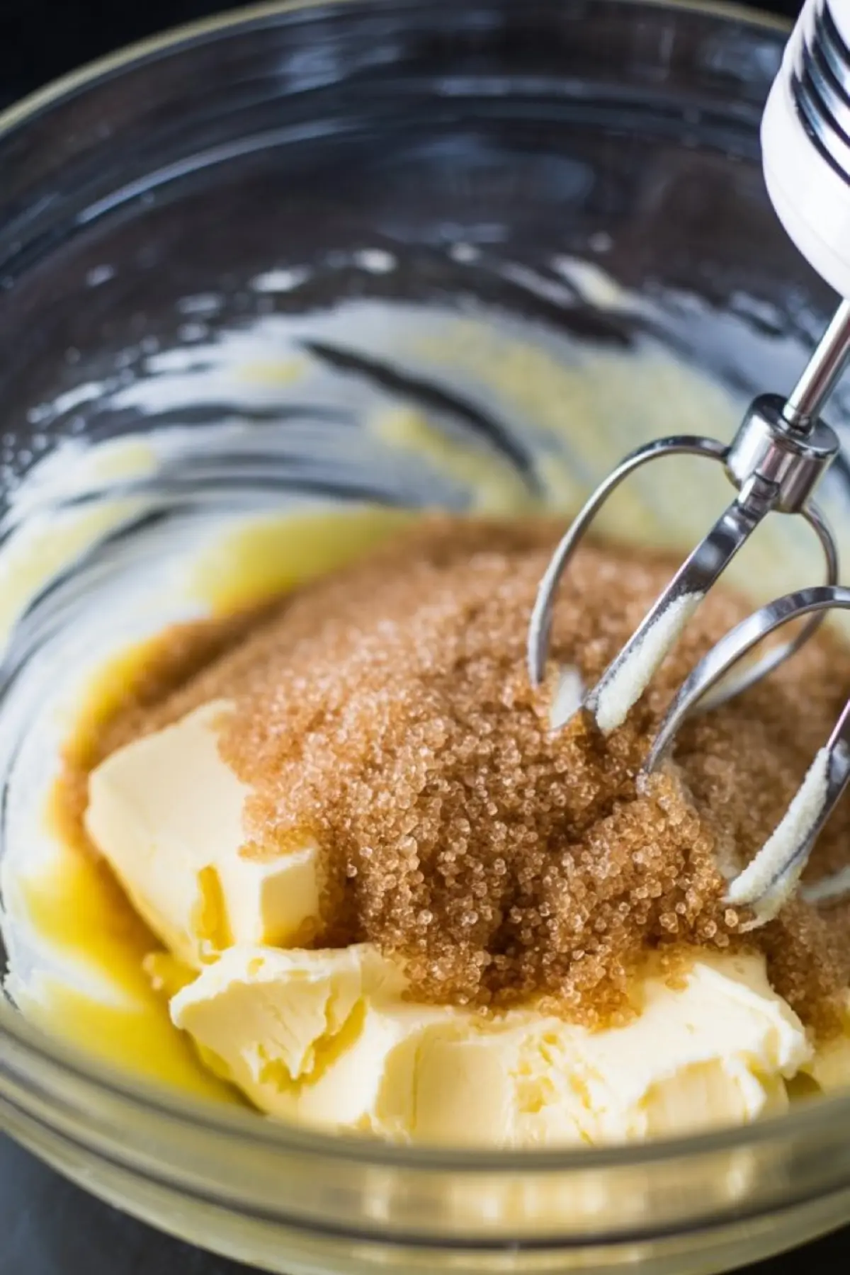 Close-up of brown sugar and softened butter in a glass bowl being creamed together with an electric hand mixer, highlighting the start of cookie dough frosting preparation.