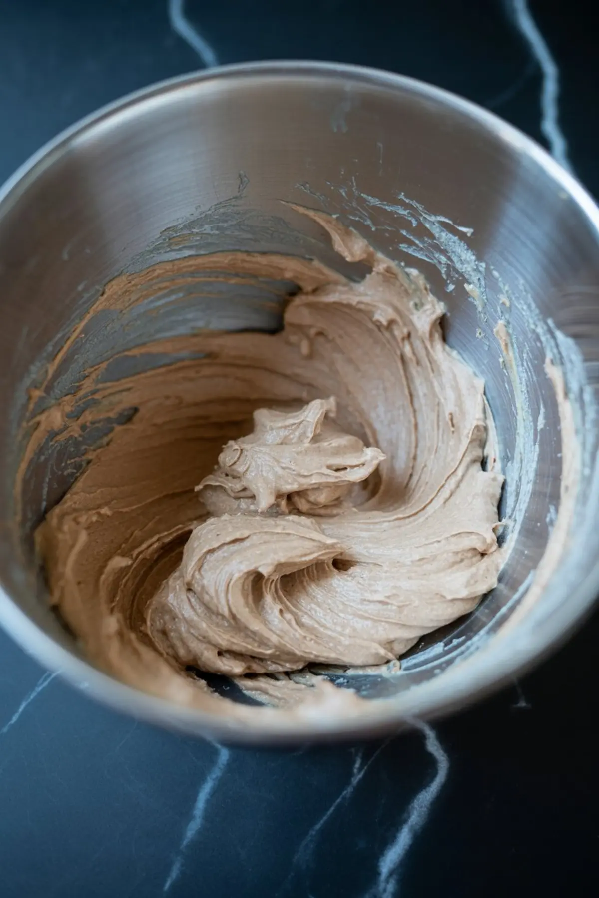 Chocolate cookie frosting batter swirled smoothly in a stainless steel mixing bowl on a dark marble countertop.
