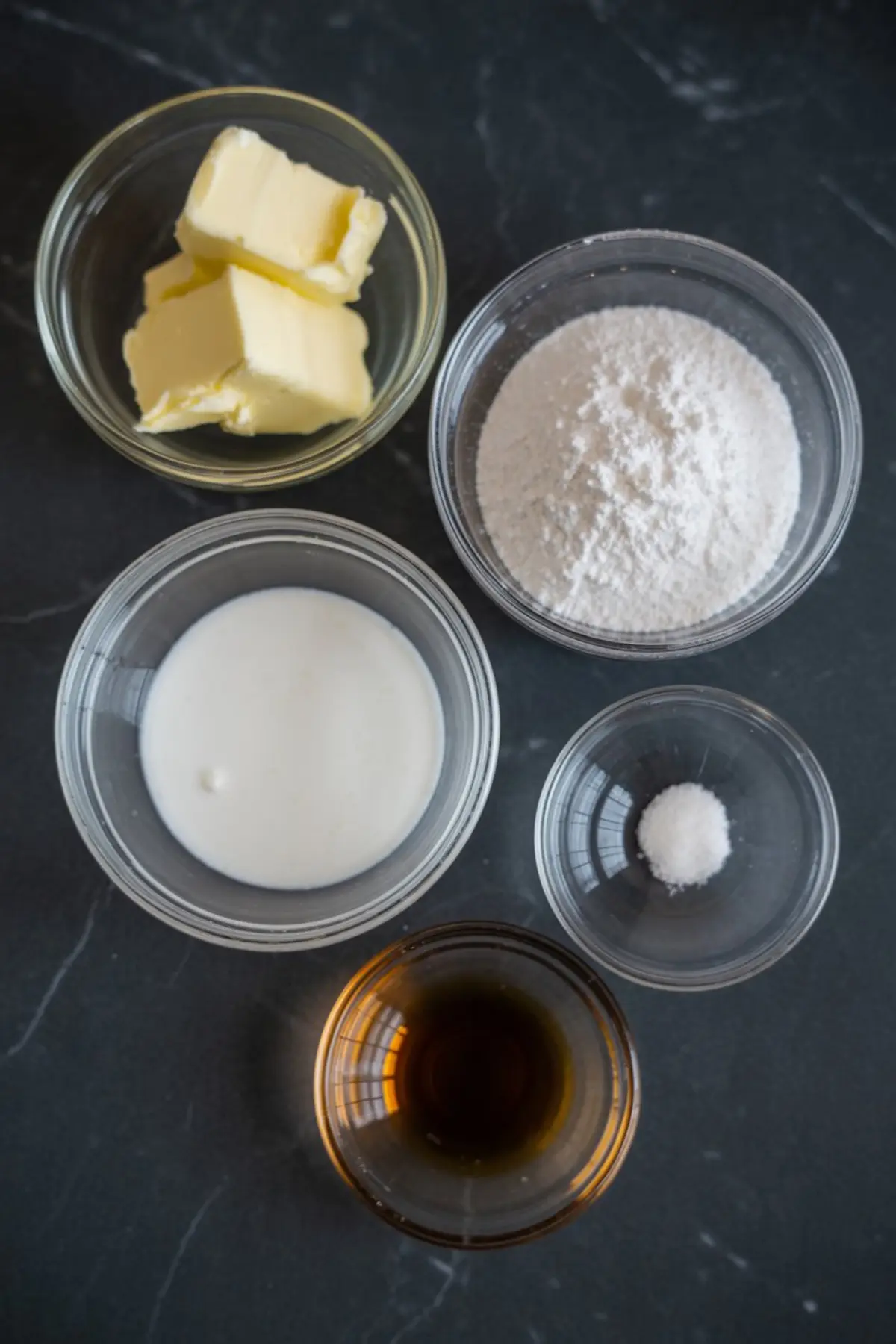 Five small glass bowls display butter, powdered sugar, cream, salt, and vanilla extract as ingredients for cookie frosting on a dark countertop.
