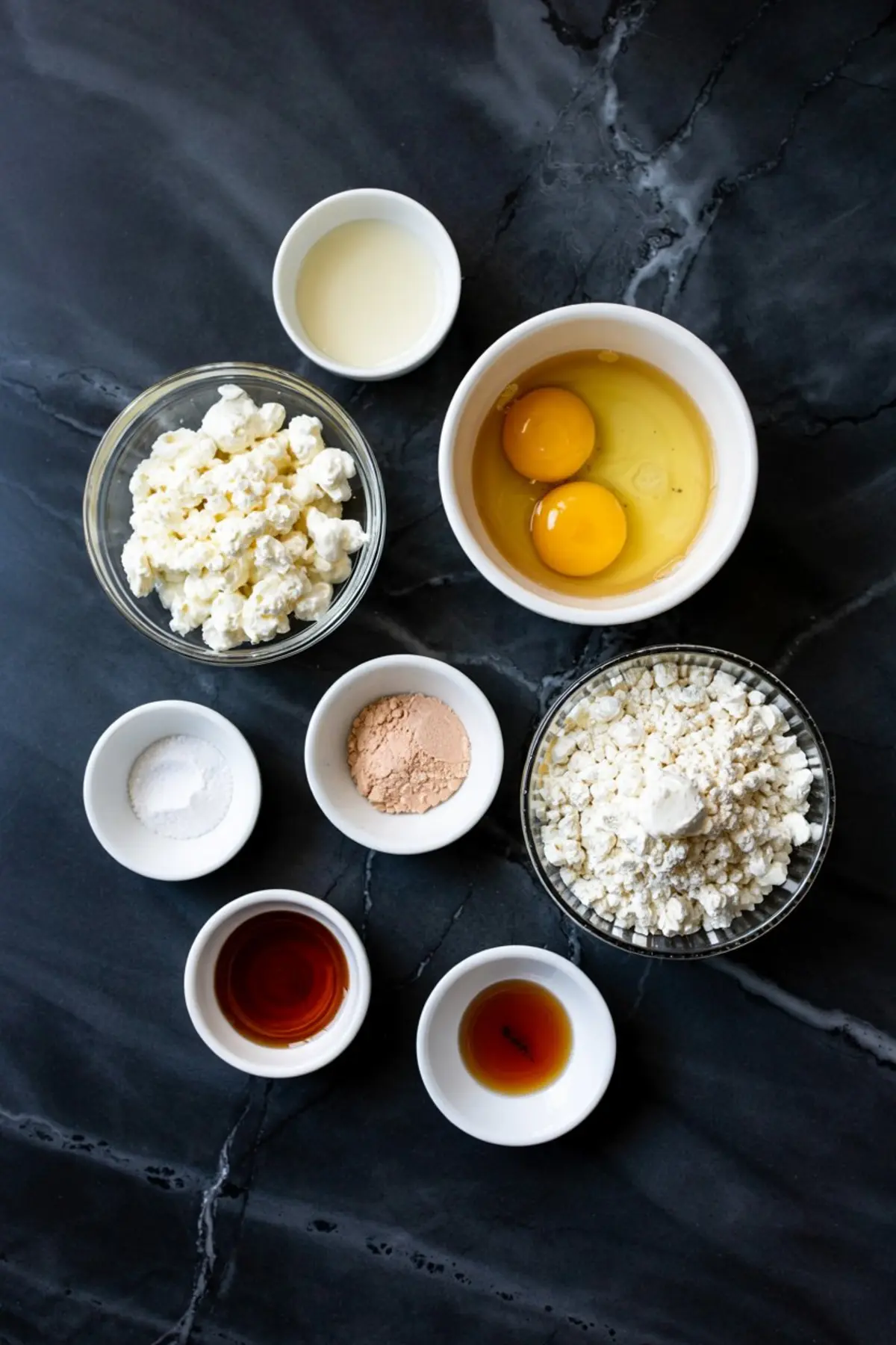 Flat lay of pancake ingredients on a black marble surface, including cottage cheese, eggs, flour, vanilla extract, baking powder, sweetener, and milk arranged in small bowls.
