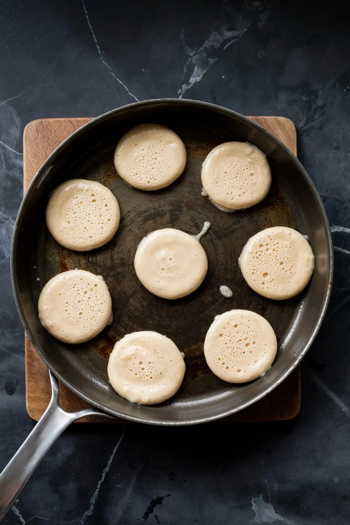 Mini pancakes cooking on a non-stick skillet, with visible bubbles forming on top of the batter, arranged in a circular pattern on a dark marble countertop.
