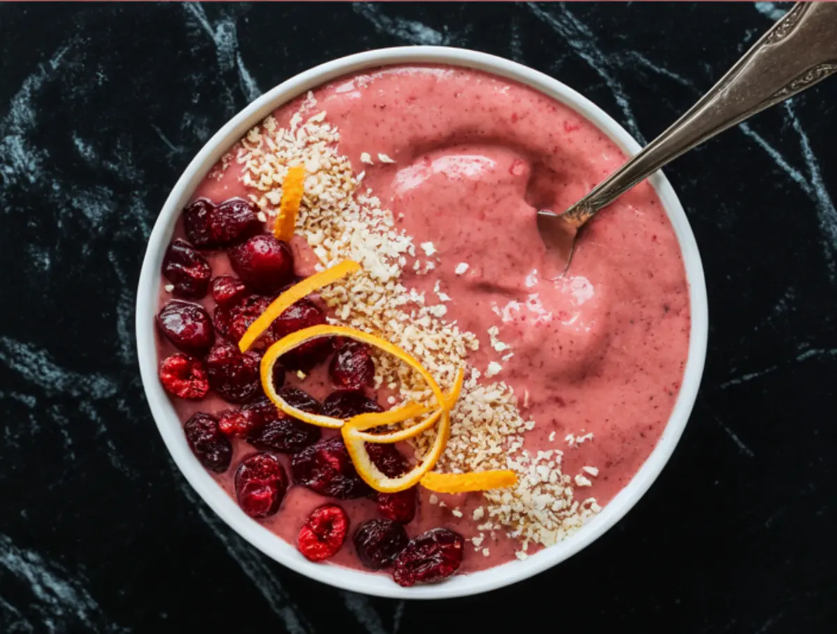 Close-up of cranberry orange smoothie bowl garnished with shredded coconut, frozen cranberries, and curly orange zest strips, served with a vintage silver spoon.