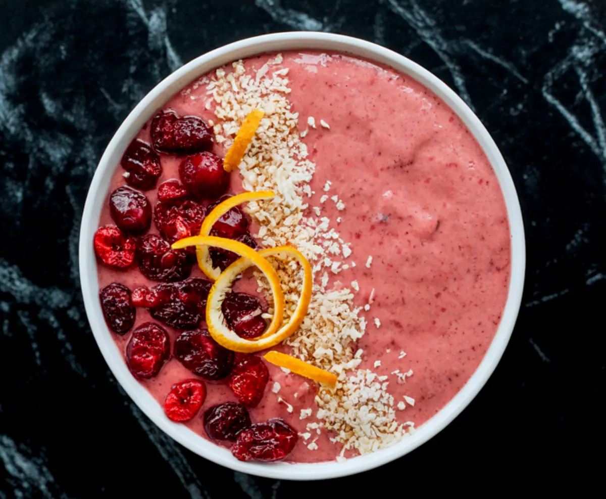 Top view of cranberry orange smoothie bowl with a row of frozen cranberries, shredded coconut, and orange zest curls, set against a black marble surface.