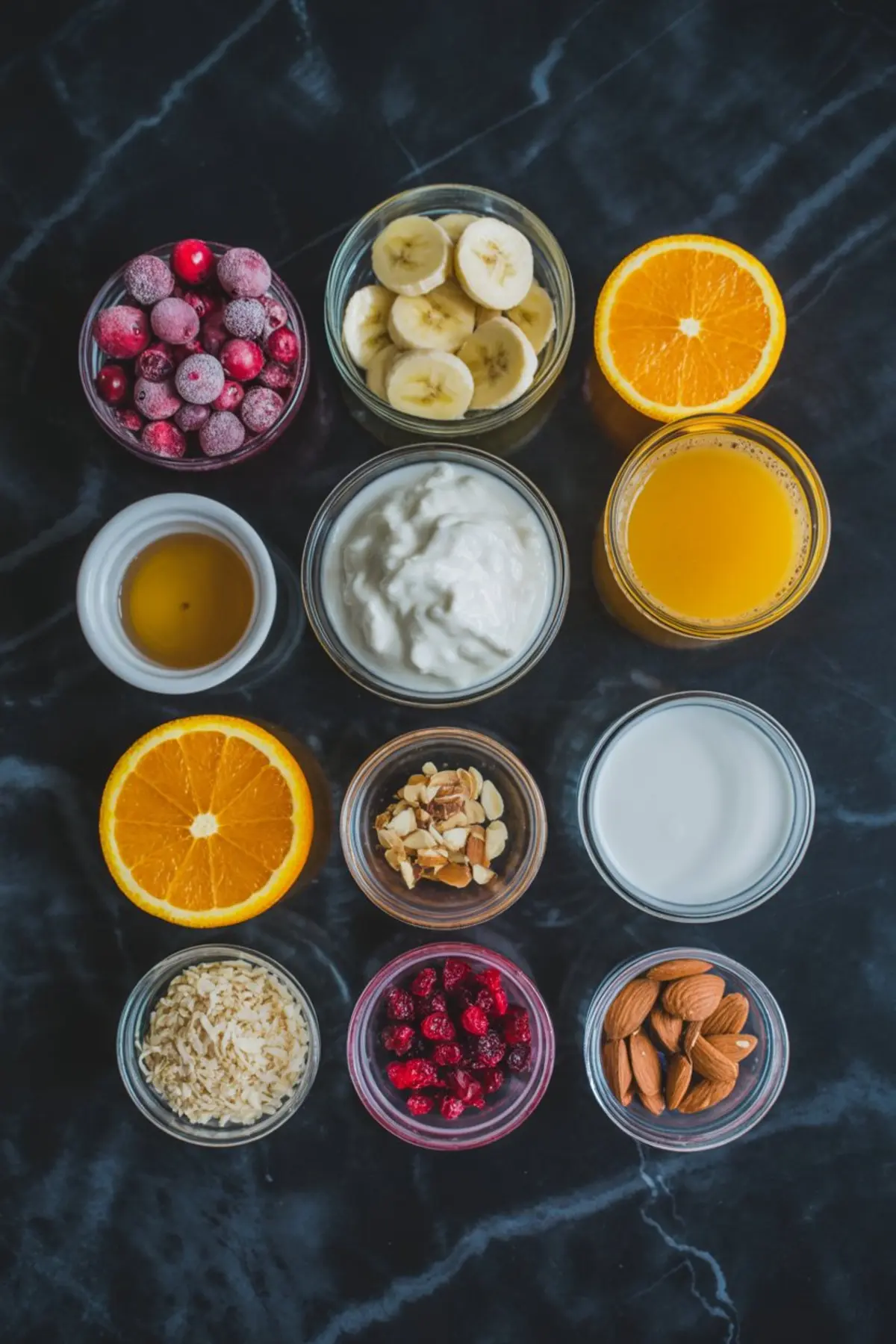 Flat lay of smoothie bowl ingredients including frozen cranberries, banana slices, orange halves, orange juice, yogurt, coconut flakes, sliced almonds, dried cranberries, almond milk, and honey arranged in small glass bowls.