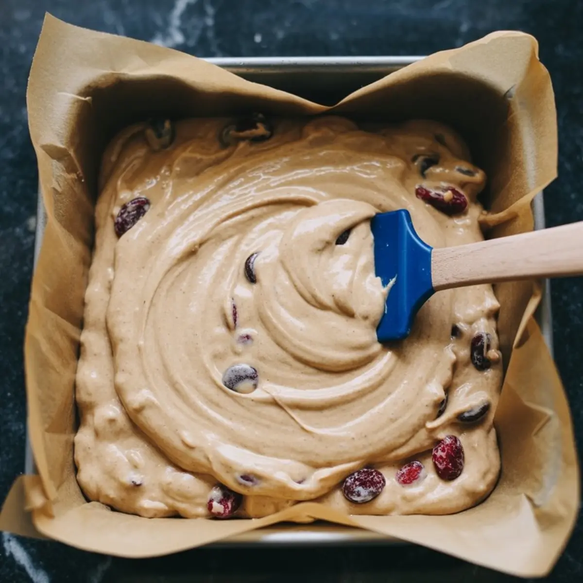 Cranberry white chocolate blondie batter swirled in a parchment-lined square pan, blue silicone spatula creating creamy ribbons on a dark marble counter.