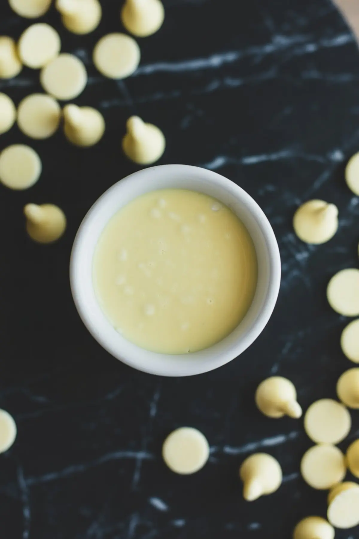Velvety melted white chocolate in a white ramekin, scattered white chocolate chips dotting a glossy black marble backdrop.