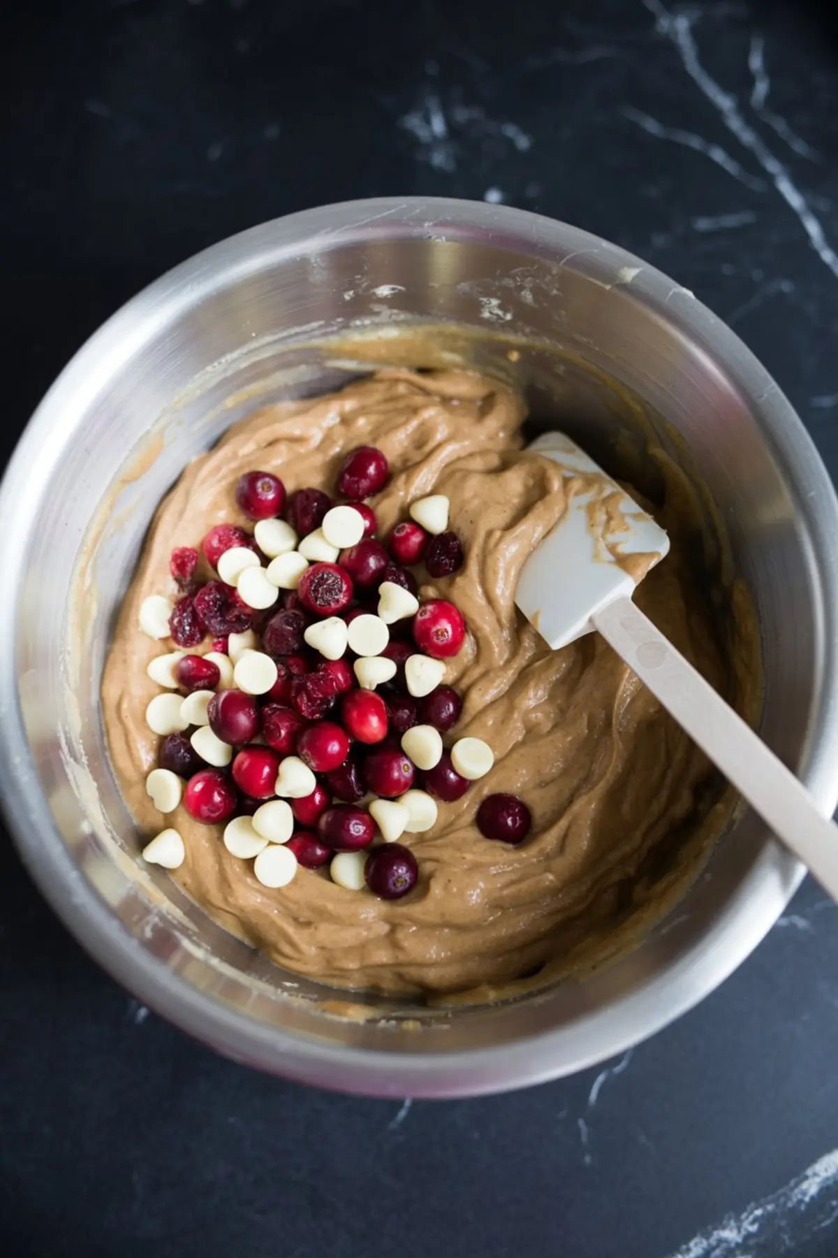 Thick blondie dough in a metal mixing bowl topped with fresh cranberries and white chocolate chips, white spatula poised to fold the mix-ins.