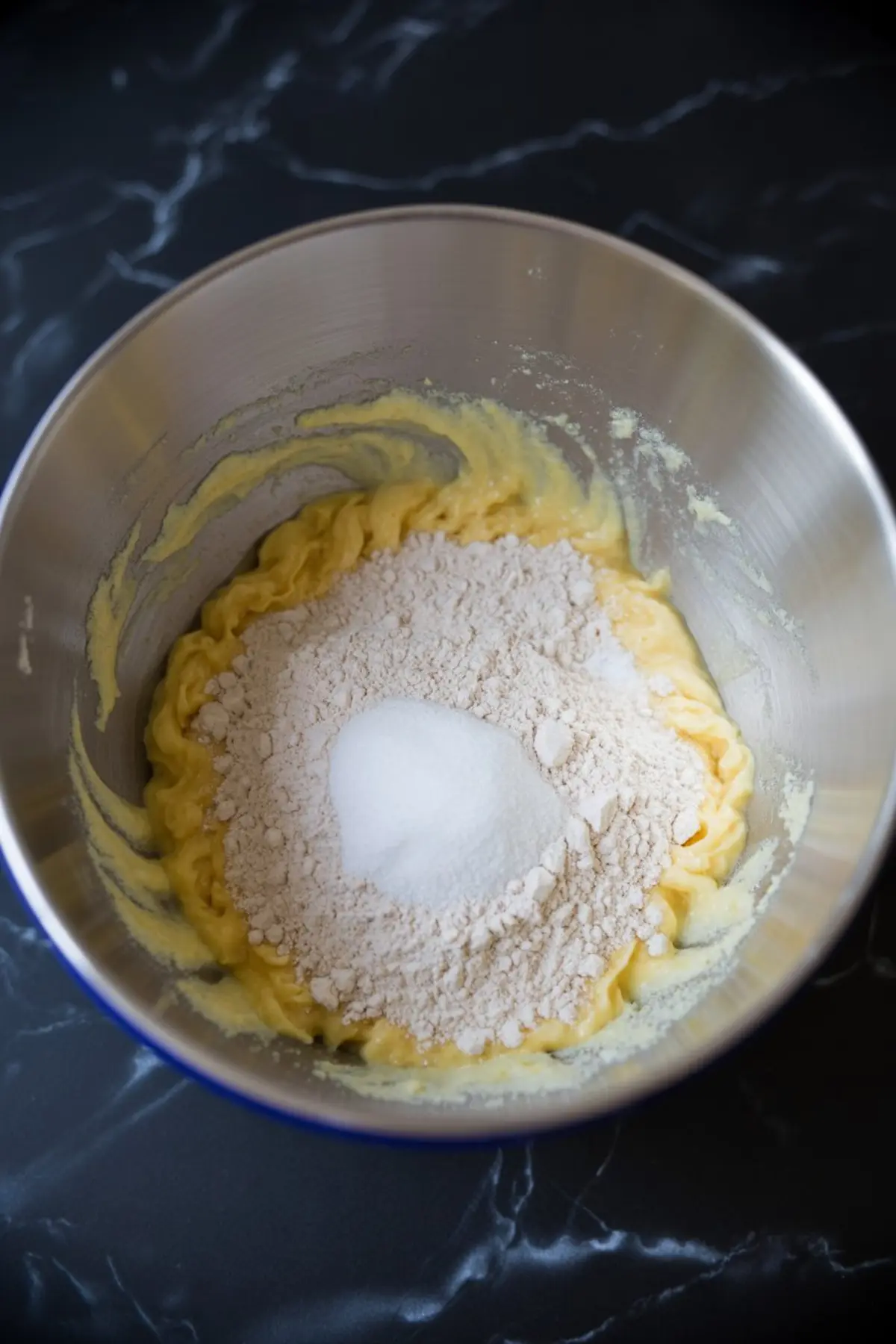 Butter-egg mixture in a steel bowl capped by a mound of flour and granulated sugar, step for combining dry ingredients in blondie batter.