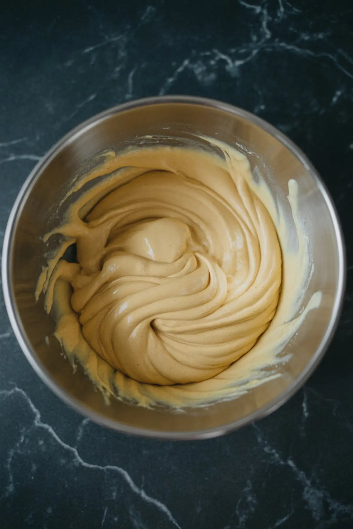 Thick golden cake batter swirled smoothly in a stainless steel mixing bowl on a black marble countertop.