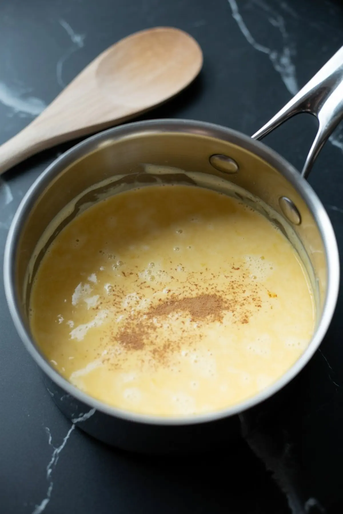 Custard mixture simmering in a stainless steel saucepan with a sprinkle of ground cinnamon and a wooden spoon nearby on a black marble counter.