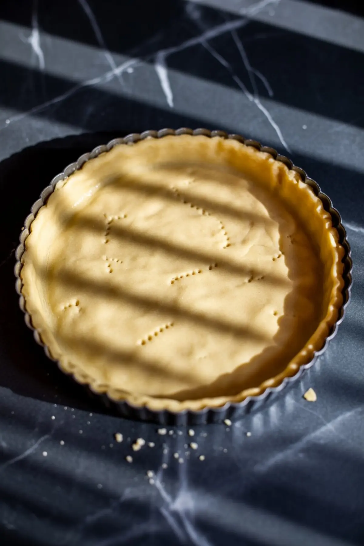 Unbaked tart crust pressed into a fluted tart pan with fork marks across the base, placed on a dark countertop with streaks of sunlight.