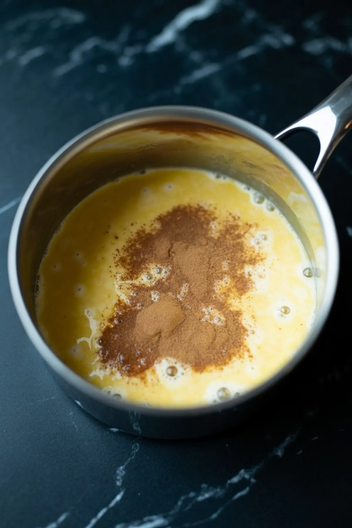 Eggnog custard mixture with cinnamon powder on top inside a stainless steel saucepan, placed on a black marble surface.