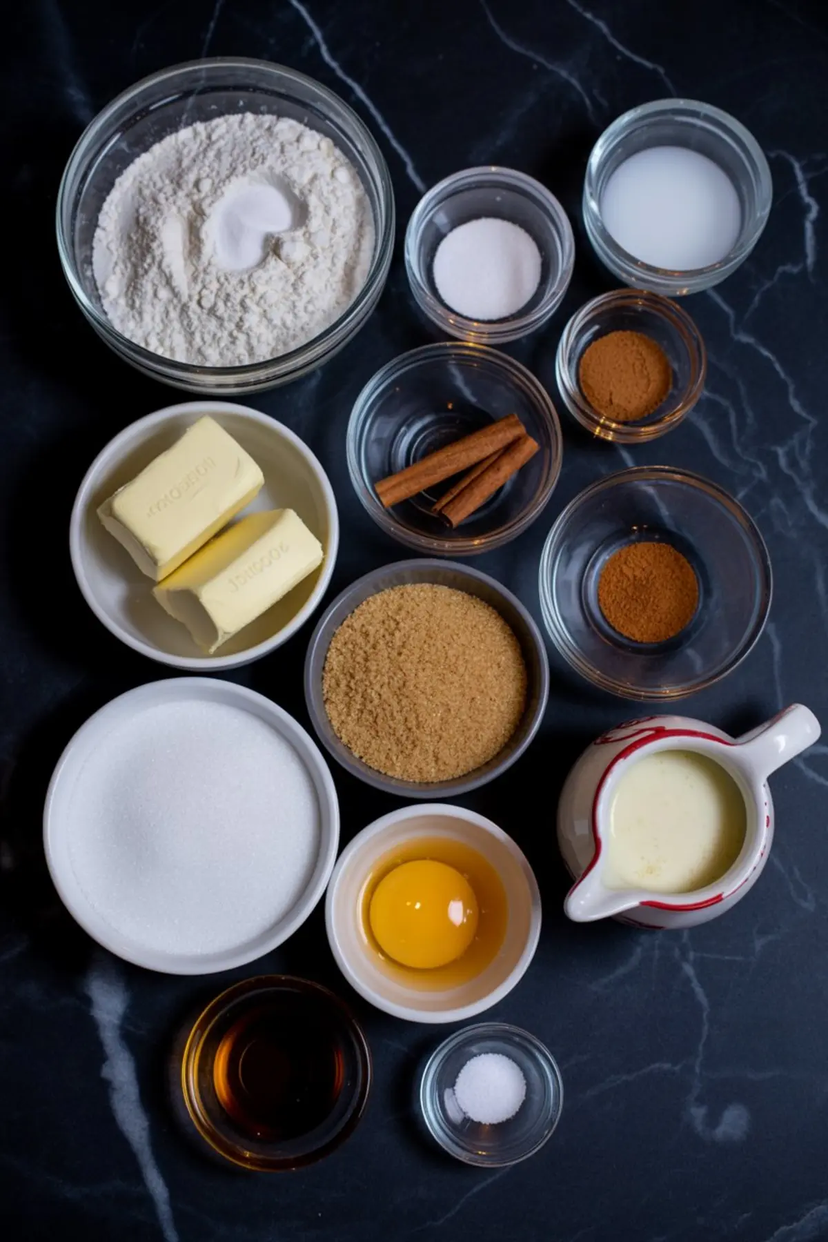Flat lay of baking ingredients on a black marble countertop including flour, butter, sugars, egg yolk, vanilla extract, cinnamon sticks, ground cinnamon, milk, and baking powder for making snickerdoodle cookies.