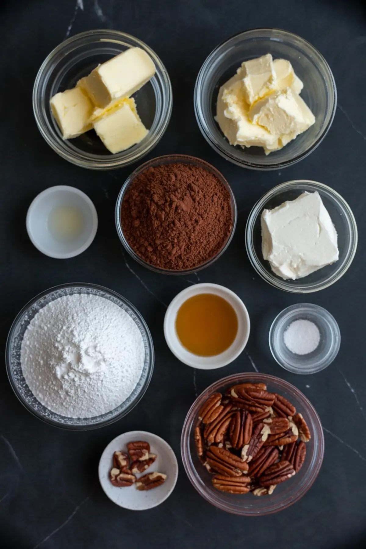 Flat lay of baking ingredients in glass bowls including cubed butter, cream cheese, cocoa powder, vanilla extract, powdered sugar, pecans, and salt on a dark slate background for German chocolate frosting.