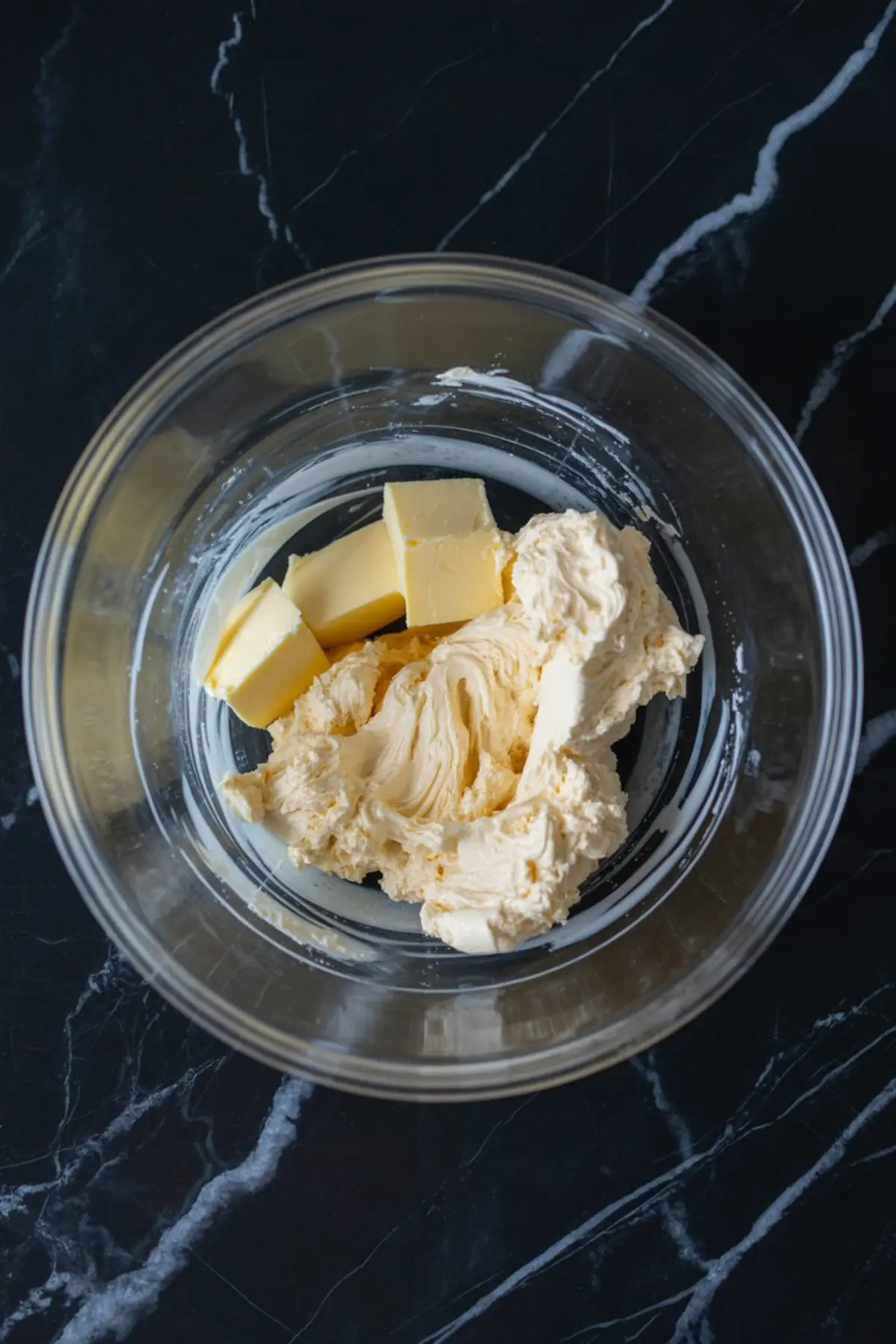 Glass mixing bowl with creamed butter and cream cheese partially blended together, set on a dark marble surface during early steps of chocolate frosting preparation.