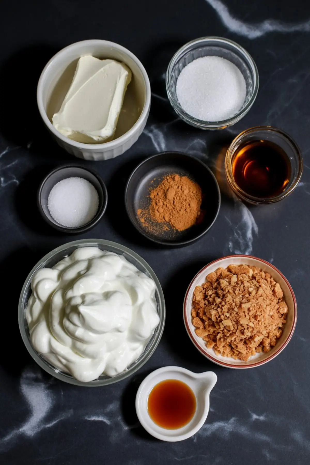 Overhead view of gingerbread cheesecake parfait ingredients arranged on a black marble surface, including whipped cream, cream cheese, sugar, cinnamon, vanilla extract, crushed cookies, and Greek yogurt.