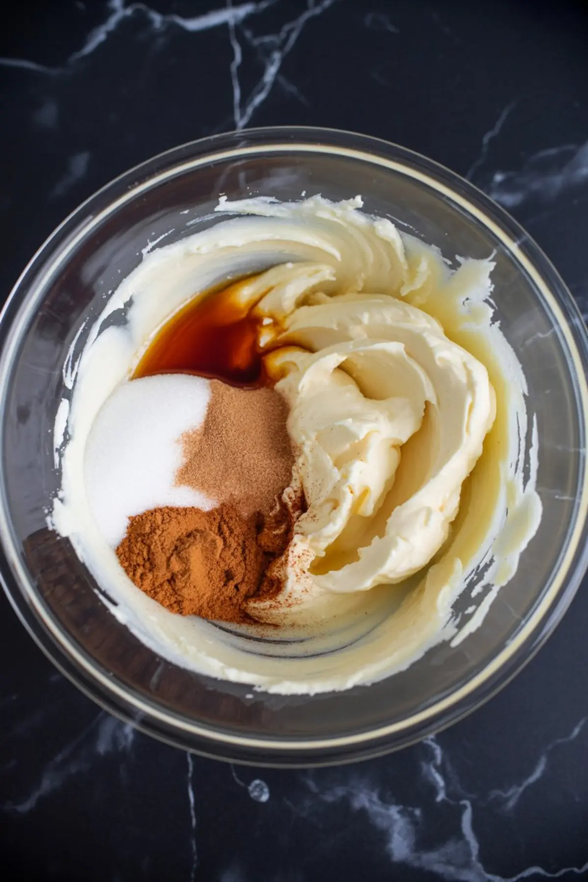 Cream cheese mixture partially combined with sugar, cinnamon, and vanilla extract in a clear glass bowl, showing rich swirls and warm spice colors.