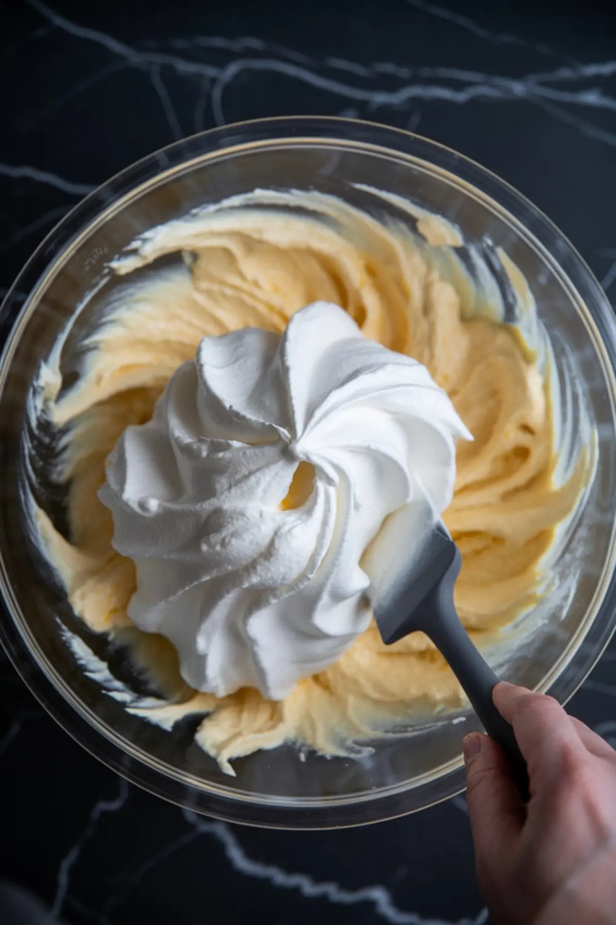 Spatula folding whipped cream into a creamy cheesecake filling in a glass mixing bowl, creating a light, airy texture for dessert preparation.