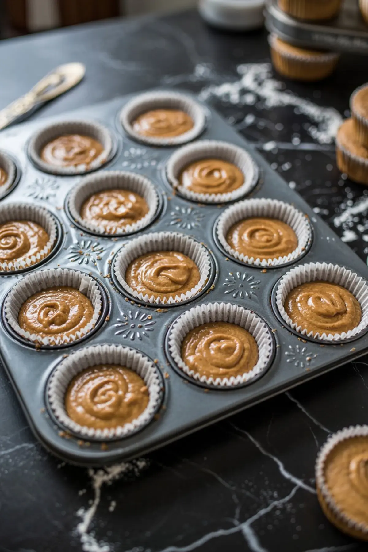 Cupcake tray lined with white paper liners filled with raw gingerbread batter, ready for baking, resting on a dark marble surface lightly dusted with flour.