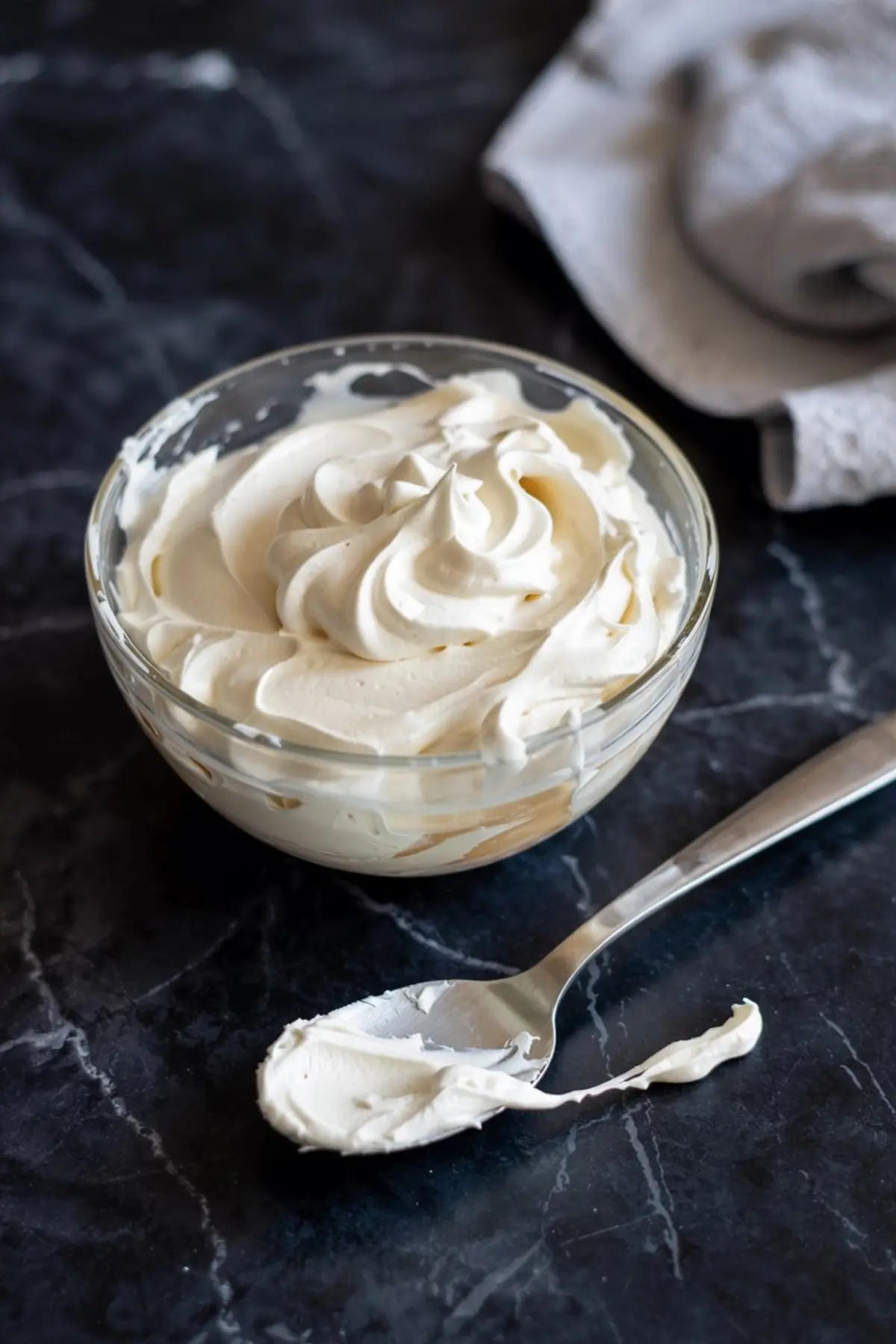 Glass bowl filled with whipped cream cheese frosting, sitting on a dark marble surface with a spoonful of frosting in front, ideal for decorating cupcakes or cakes.