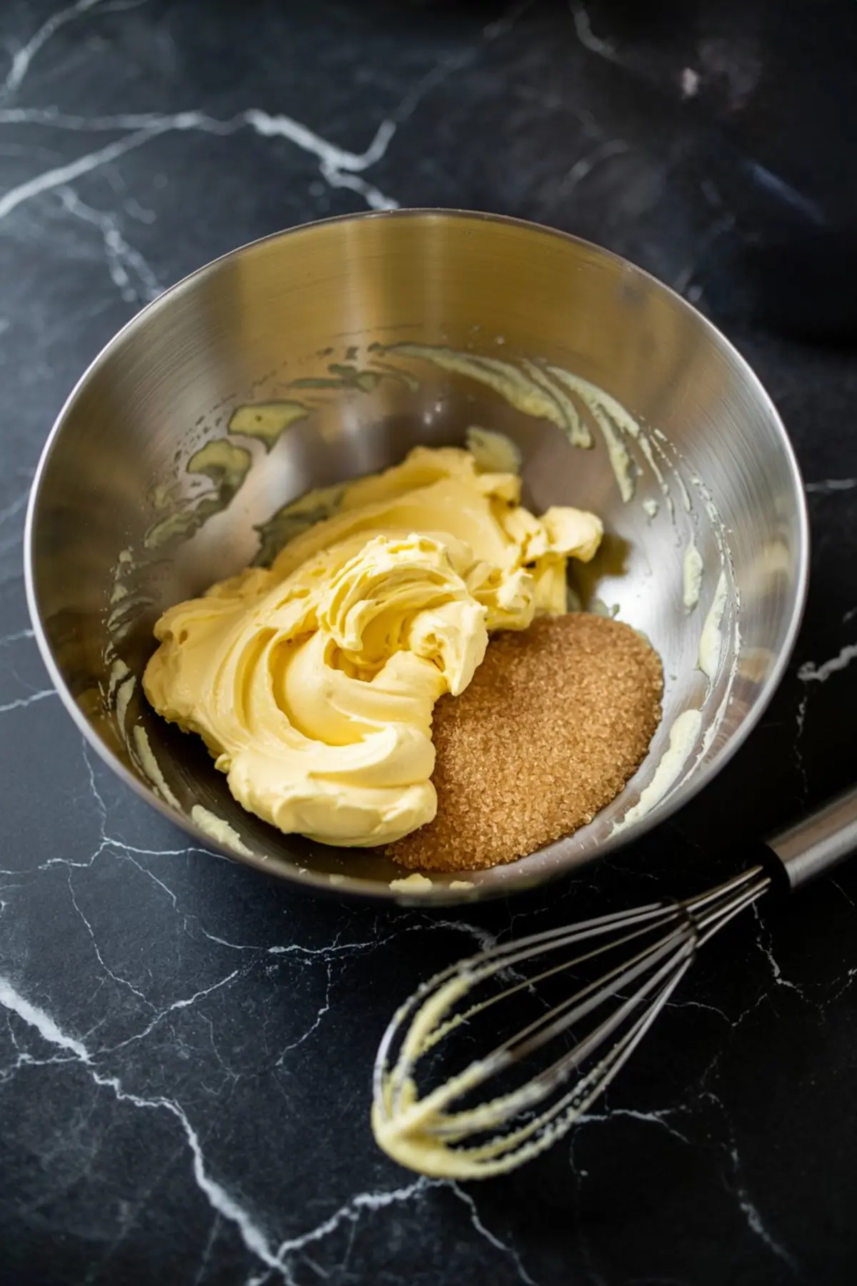 Stainless steel mixing bowl containing softened butter and brown sugar, placed on a black marble counter with a whisk beside it, showing the beginning steps of cupcake batter preparation.