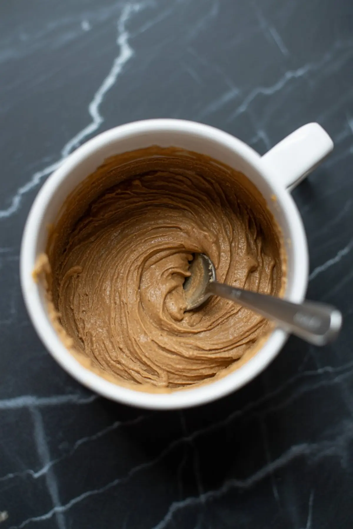 Thick gingerbread mug cake batter swirled with a spoon inside a white mug, showing a smooth, creamy consistency on a dark marble surface.