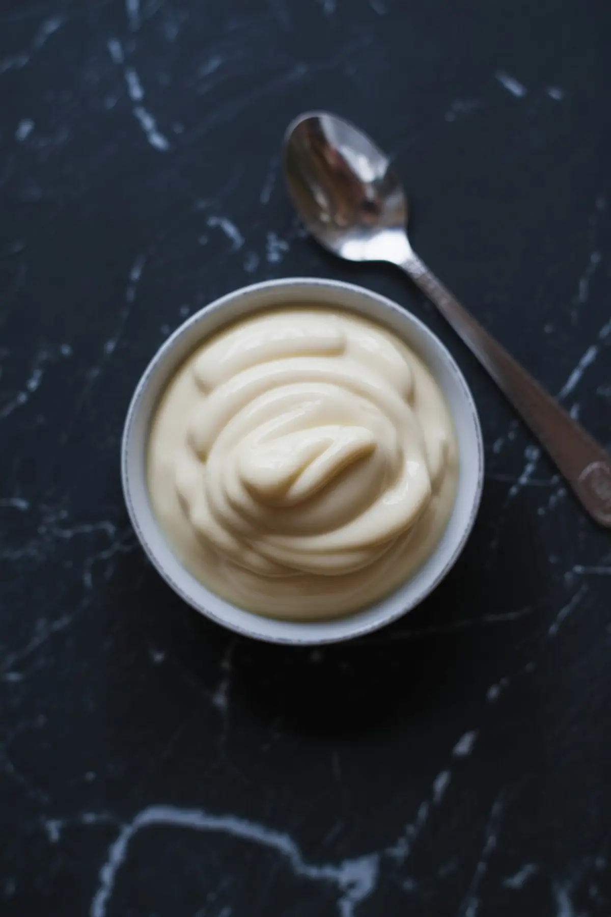 Small bowl of smooth vanilla frosting or whipped cream with a glossy texture, placed beside a silver spoon on a black marble background.