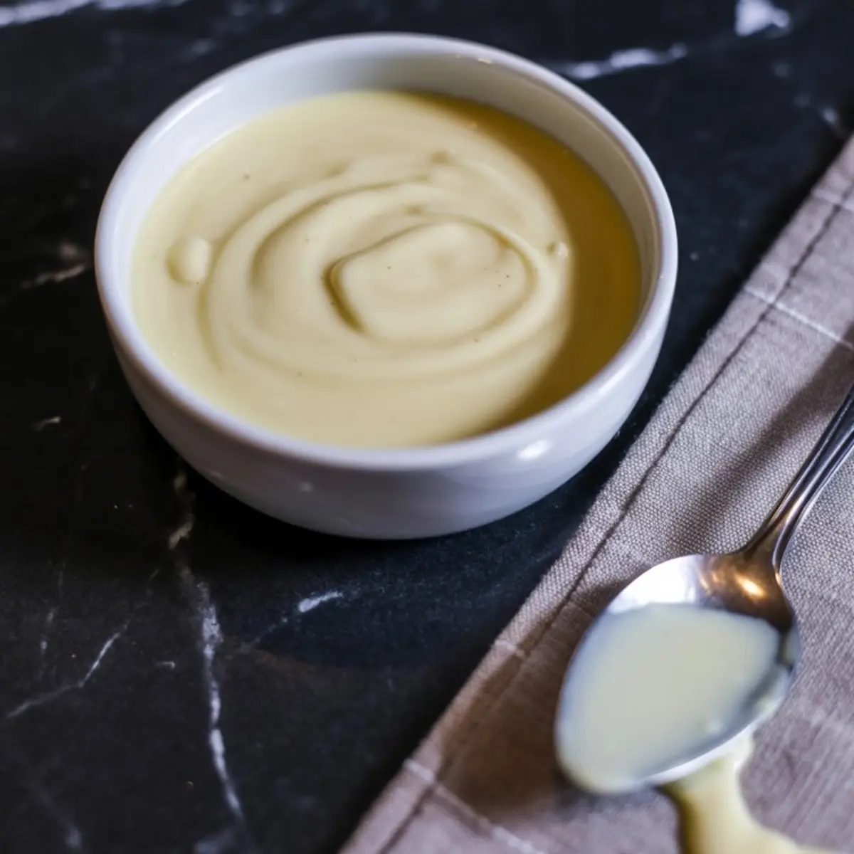 A small white bowl filled with creamy white chocolate icing on a dark countertop with a spoon resting on a beige cloth beside it.