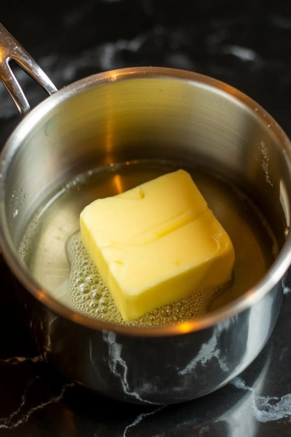 A saucepan with butter melting over heat, showing bubbling edges on a dark marble countertop.