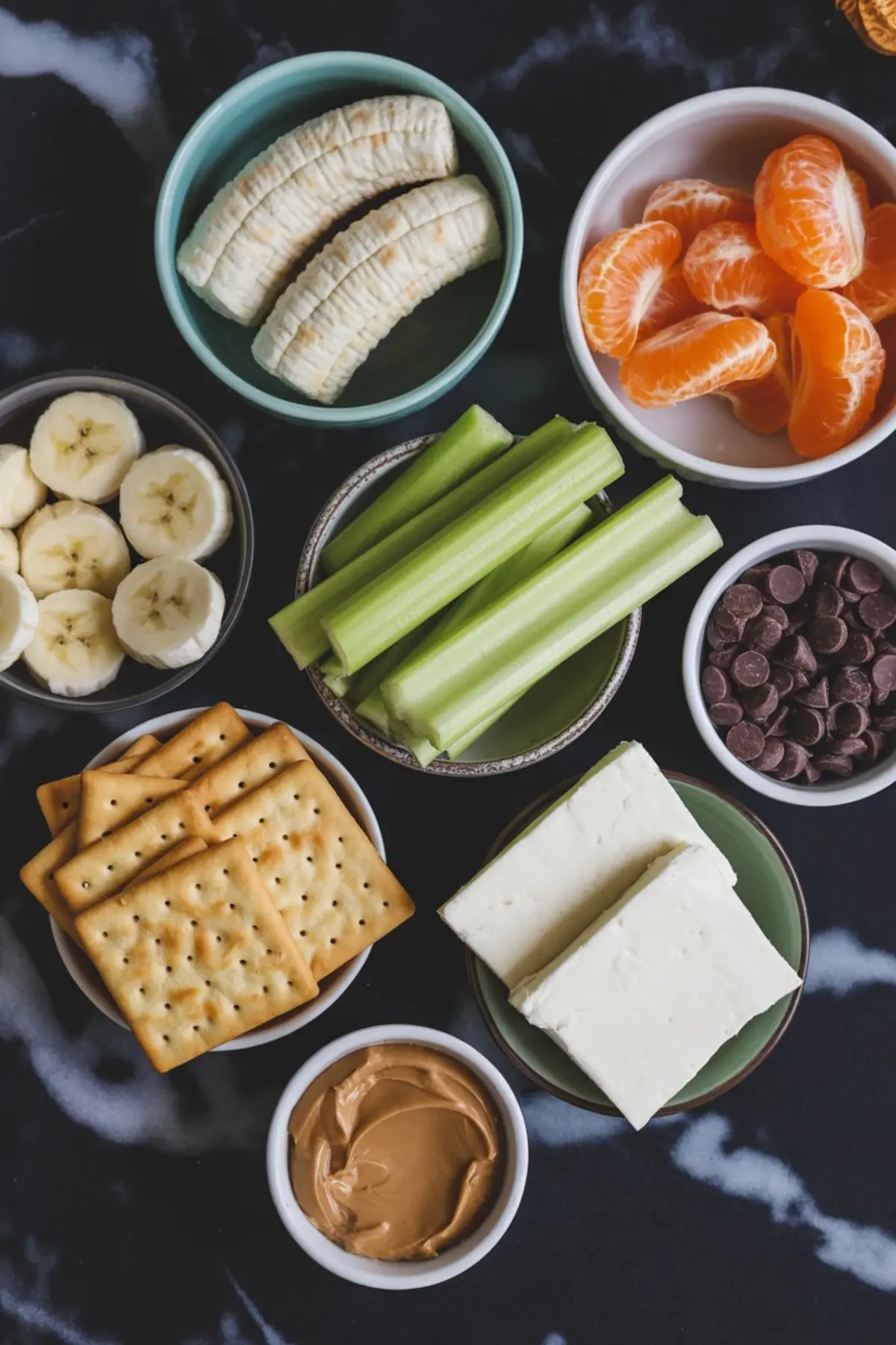 Flat lay of healthy snack ingredients including bananas, clementine oranges, celery sticks, chocolate chips, peanut butter, cream cheese, and square crackers arranged in small bowls on a dark surface.

