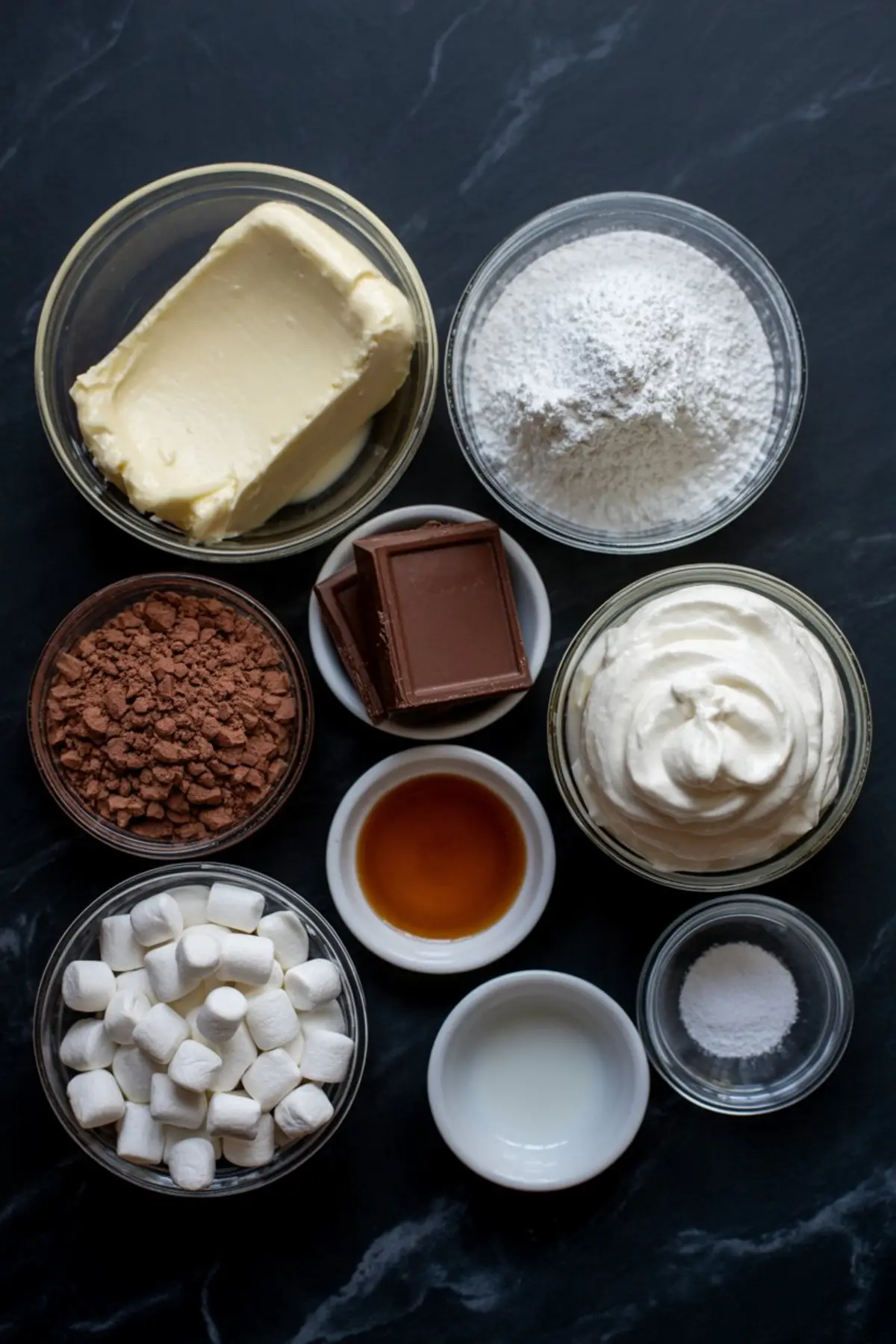 Flat lay of hot chocolate cheesecake dip ingredients in glass bowls on a black marble surface, including butter, powdered sugar, cocoa powder, chocolate squares, whipped cream, mini marshmallows, vanilla extract, milk, and salt.
