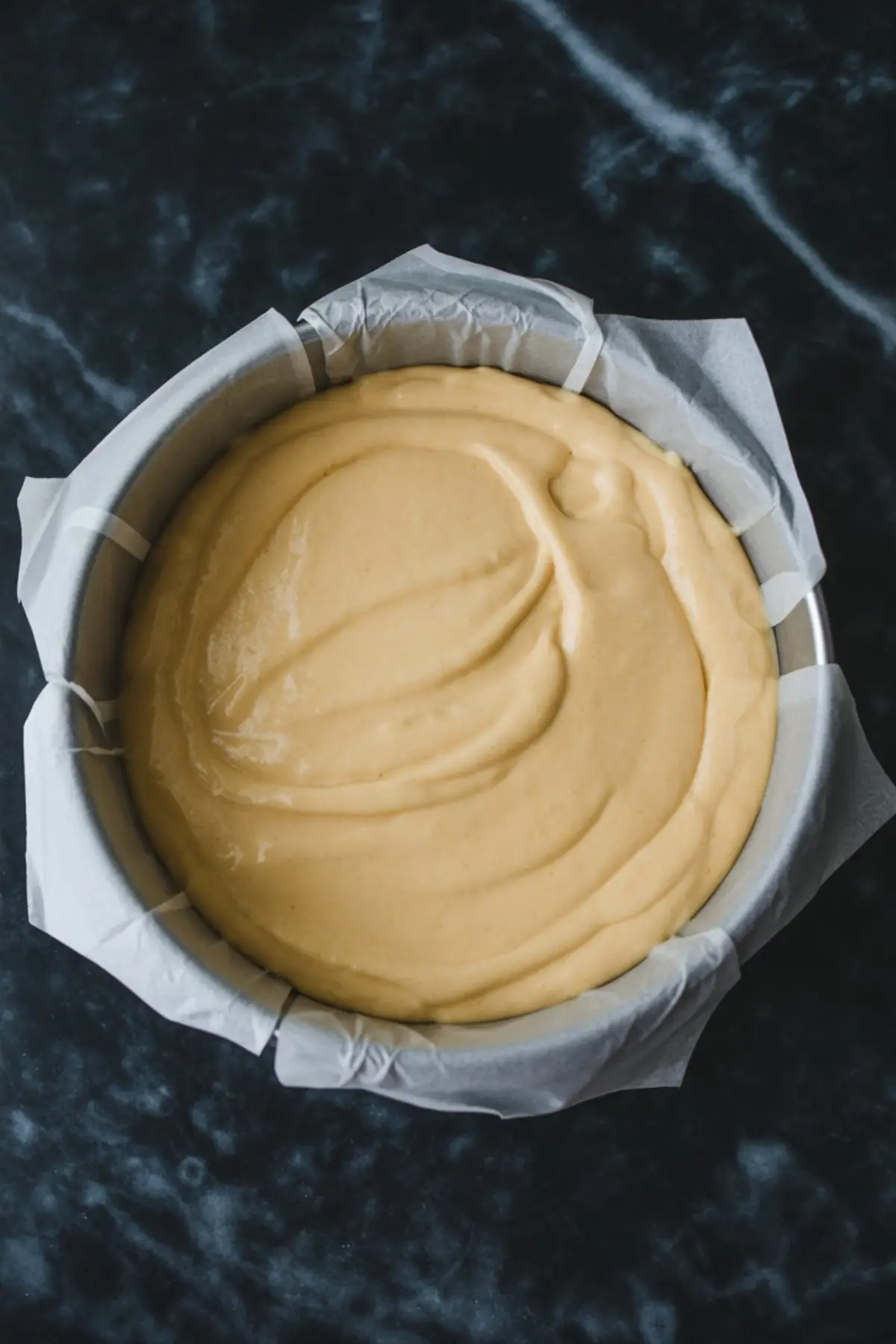 Round cake pan lined with parchment paper filled with raw Japanese cheesecake batter, smooth and evenly spread over a dark marble surface.