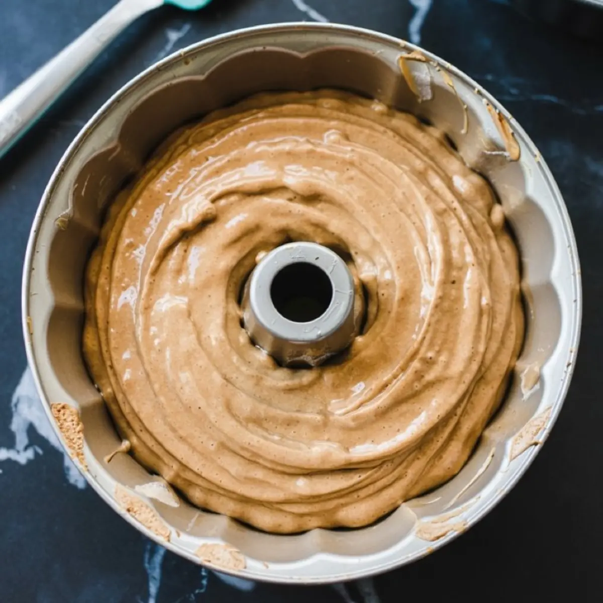 Thick maple cake batter swirled evenly in a bundt pan, ready for baking, set on a dark marble countertop with a teal spatula in view.