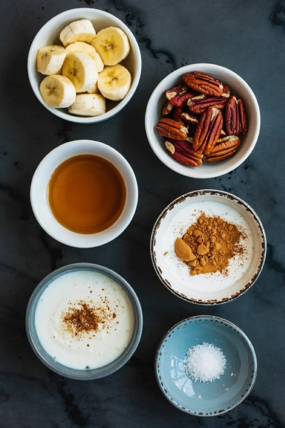 Flat lay of maple pecan smoothie bowl ingredients in small bowls, including sliced bananas, whole pecans, maple syrup, ground cinnamon, Greek yogurt with cinnamon, and sea salt on a dark stone background.
