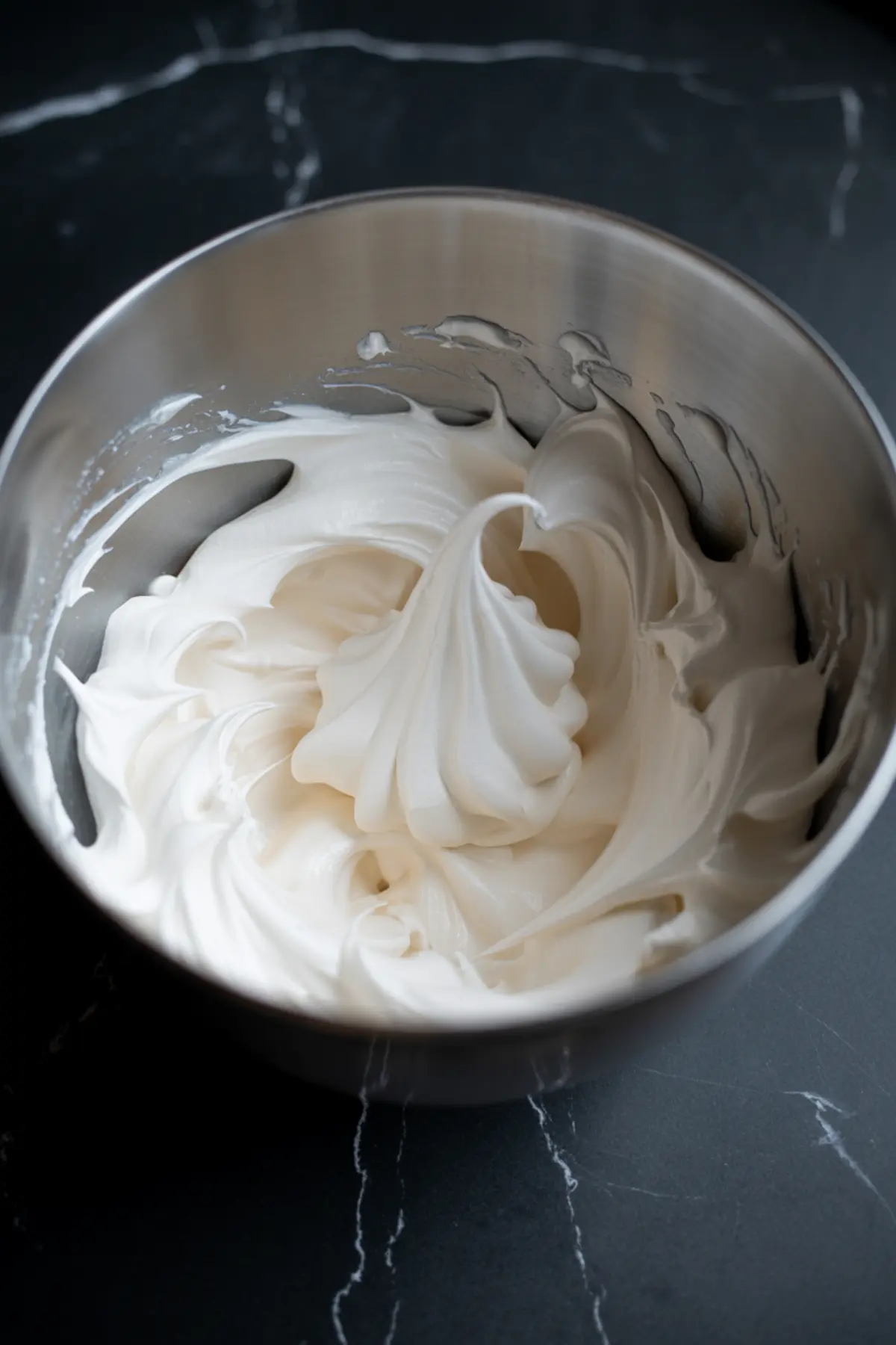 Fluffy marshmallow frosting with stiff peaks swirled in a stainless steel mixing bowl, whipped to a glossy finish for cupcakes or cakes.
