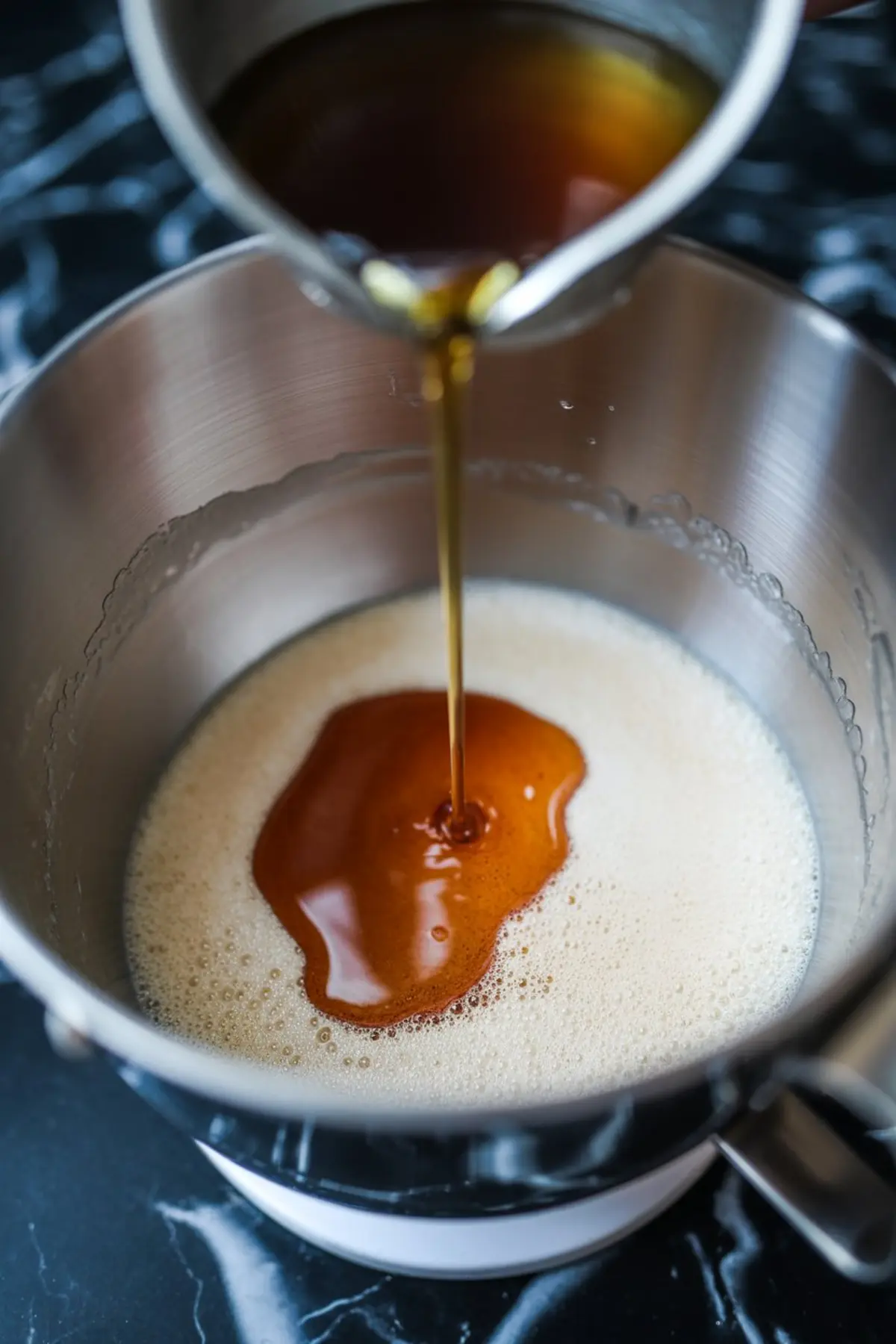 Golden syrup being poured into a mixing bowl of whipped egg whites, forming the base for smooth marshmallow frosting.
