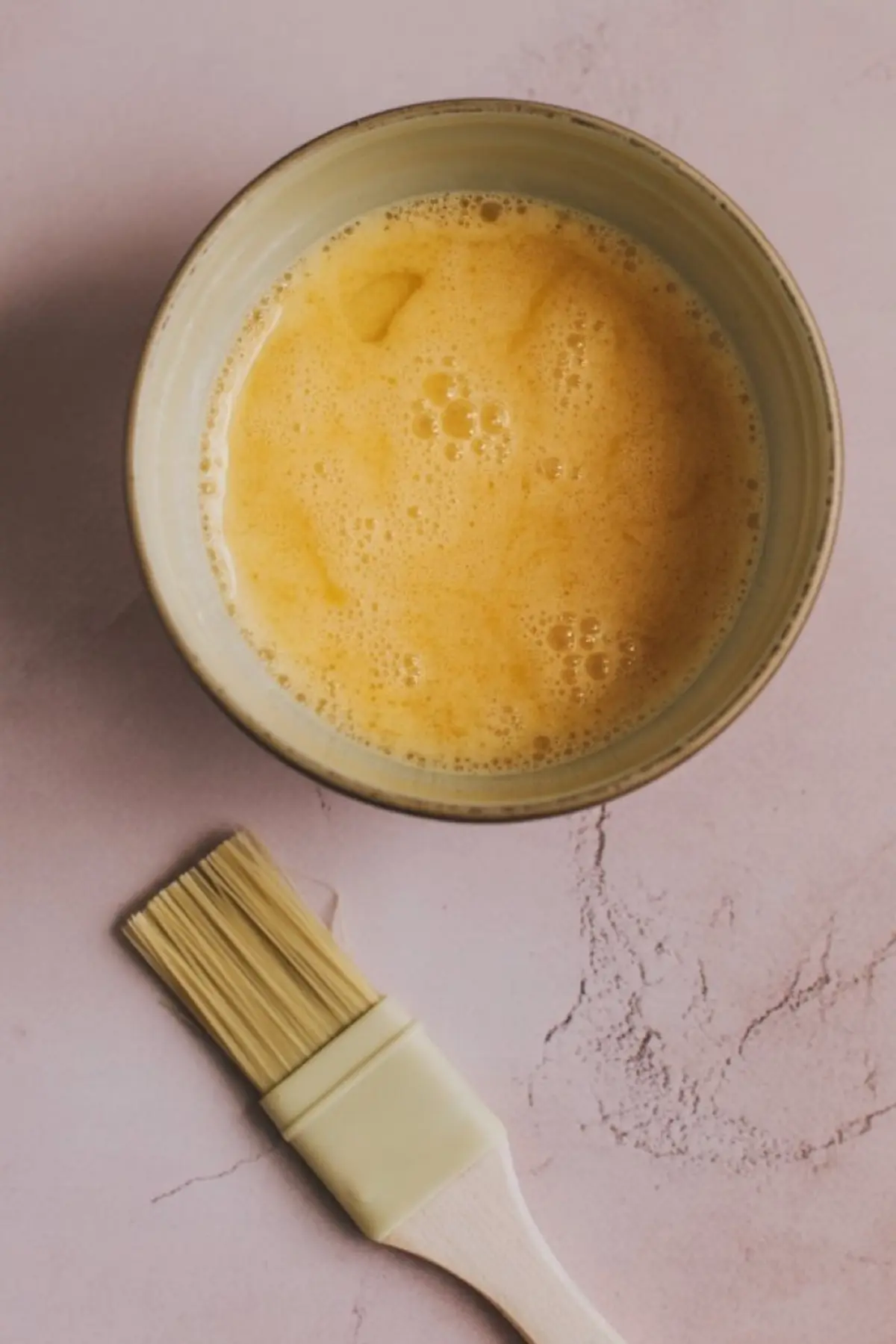 A ceramic bowl filled with whisked egg wash sits on a light pink textured surface, with a silicone pastry brush placed beside it for baking preparation.

