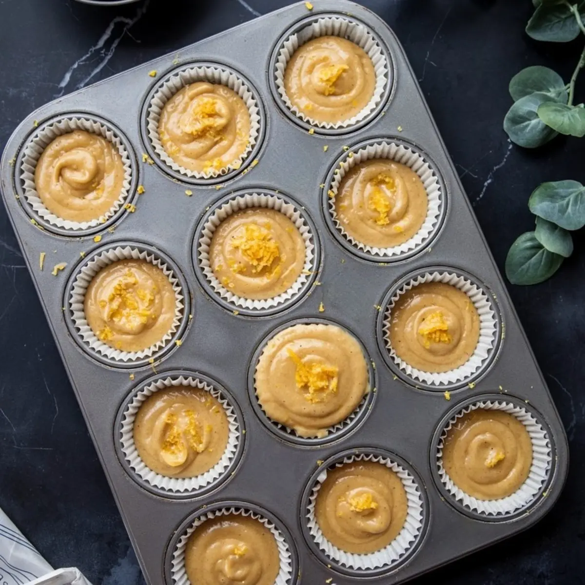 Twelve muffin liners filled with orange muffin batter topped with orange zest, arranged in a metal muffin tray on a black surface with green leaves nearby.