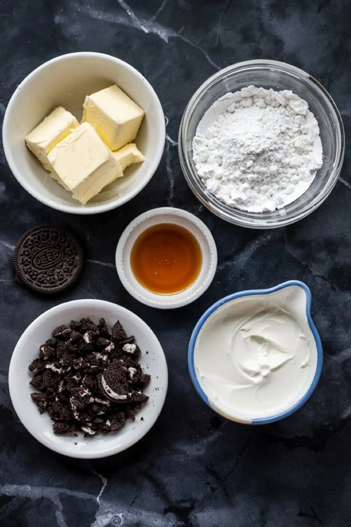 Overhead view of Oreo frosting ingredients including butter cubes, powdered sugar, vanilla extract, crushed Oreos, and cream cheese on a dark marble background.