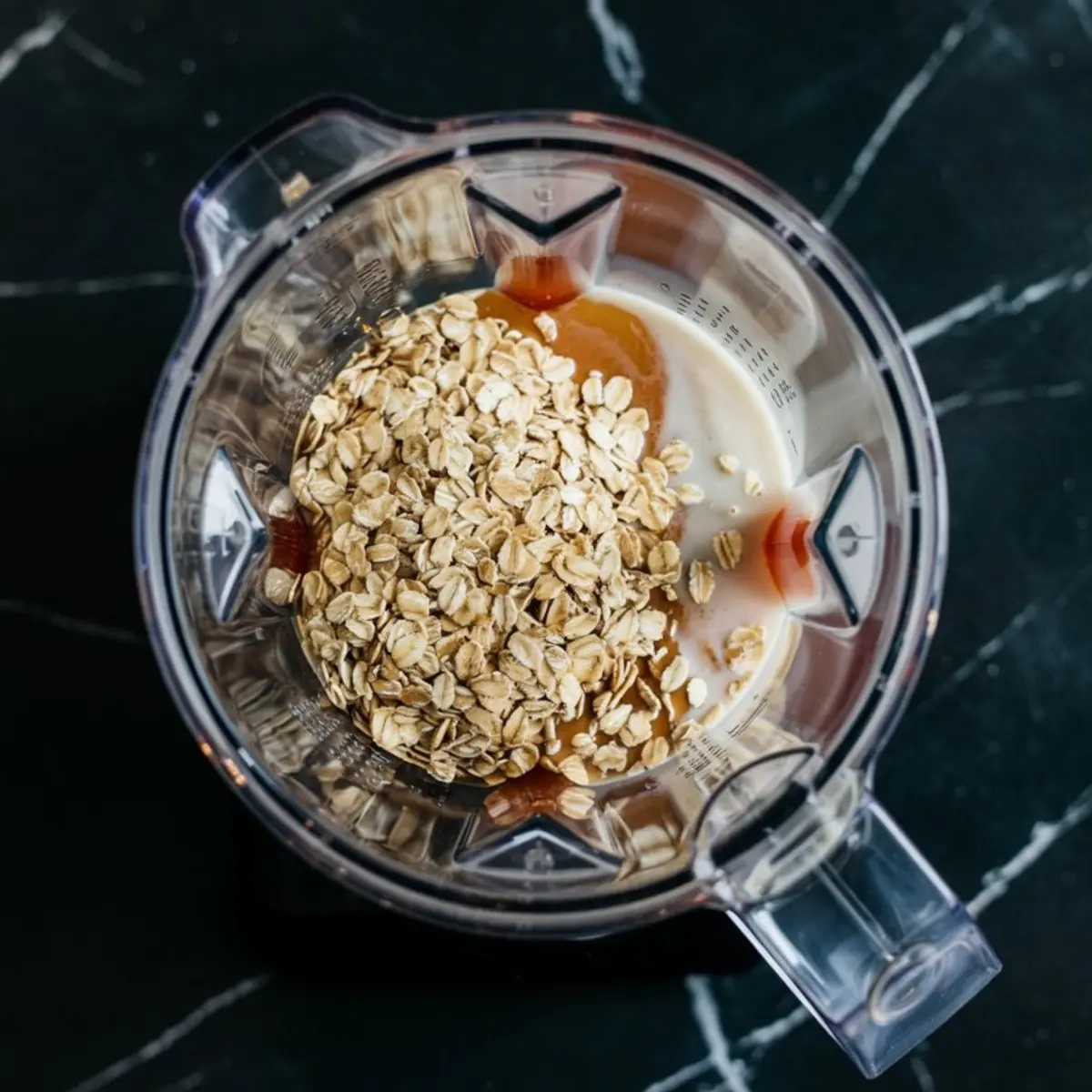 Overhead view of oats, milk, and honey in a clear blender jug ready to blend for a smoothie base.