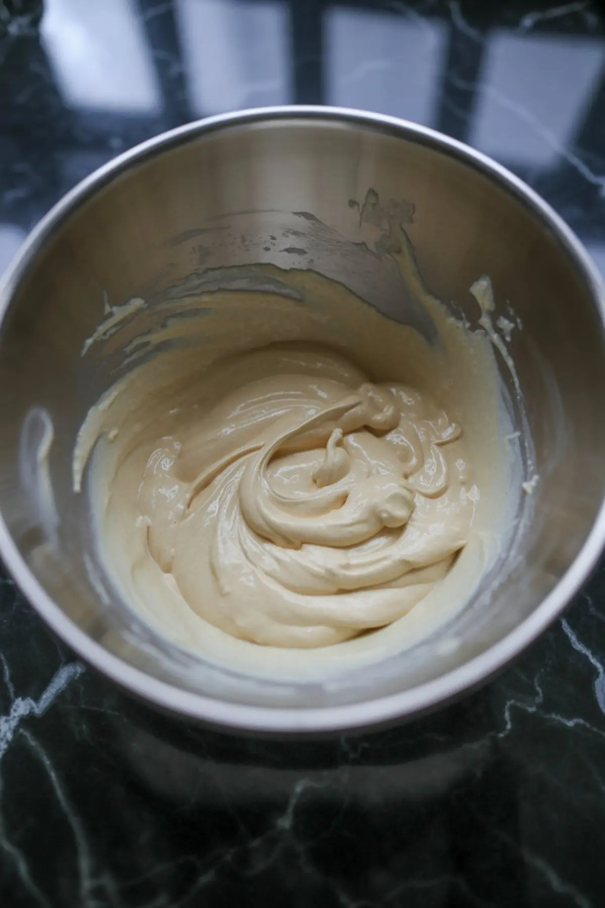 Thick vanilla cheesecake batter swirled in a stainless steel mixing bowl on a black marble countertop, showing a rich and creamy texture ready for baking.