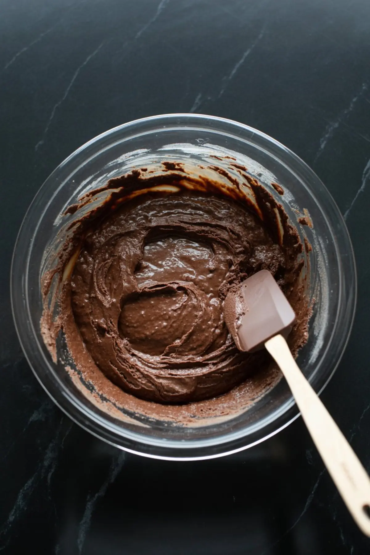 Glass bowl filled with thick chocolate brownie batter being mixed with a spatula on a dark stone surface, showing rich texture for dessert preparation.