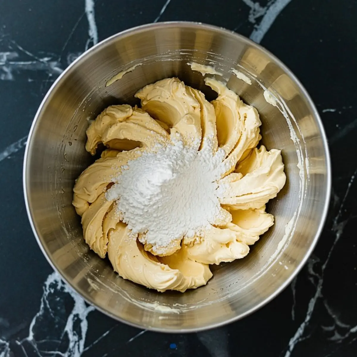 Butter and powdered sugar mixture in a stainless steel mixing bowl, prepared for a dessert base on a black marble surface.
