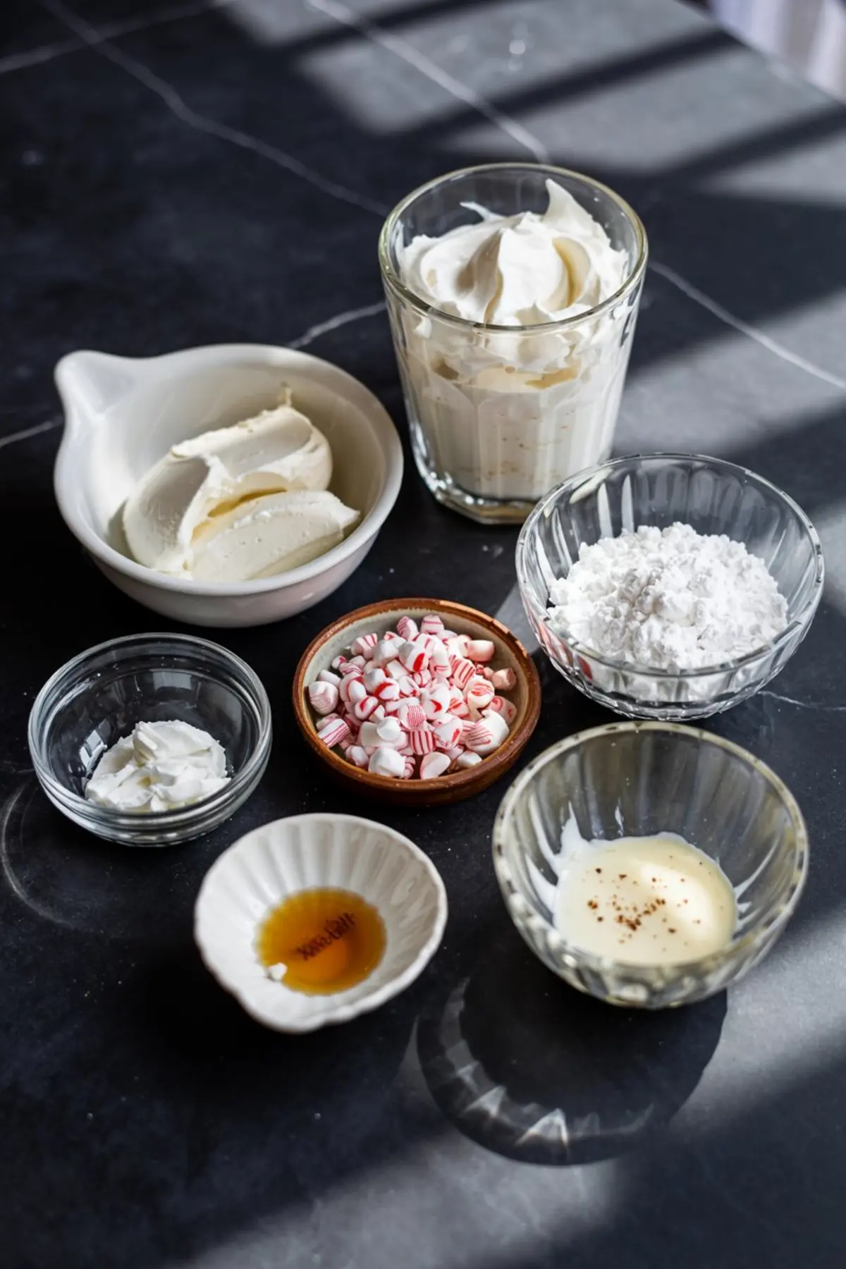 Flat lay of peppermint cheesecake dip ingredients including cream cheese, whipped topping, powdered sugar, crushed peppermint, vanilla extract, and flavoring in assorted glass and ceramic bowls.
